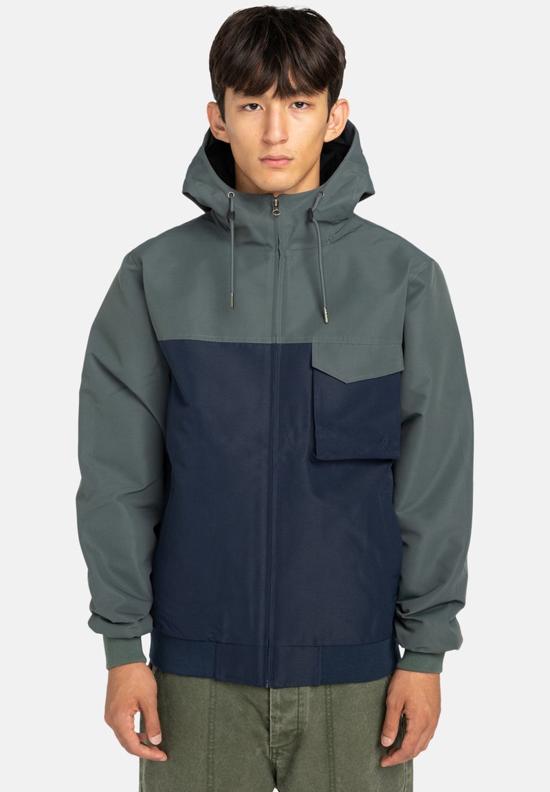 Giacca con cappuccio di colore grigio-verde nella parte superiore e blu navy nella parte inferiore. Presenta una tasca frontale, chiusura con zip e polsini elasticizzati.