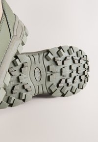 Grüne, robuste Außensohle mit tiefen, aggressiven Profilen. Strukturierter Gummi für besseren Halt. Verstärkte Kanten und ovale Markenprägung.