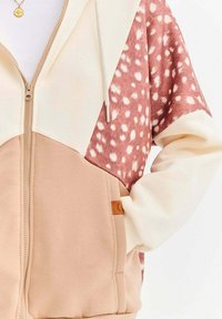 Bluza z kapturem zapinana na zamek, z pastelowymi blokami kolorów: kremowym, jasnoróżowym i brązowym, cętkowanym wzorem. Posiada oznaczoną metkę po boku.