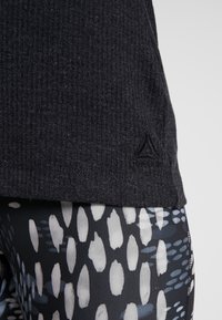 Svart ribbad topp med Reebok-logotyp vid fållen, tillsammans med mönstrade leggings med vita och blå förlängda former på en mörk bakgrund.