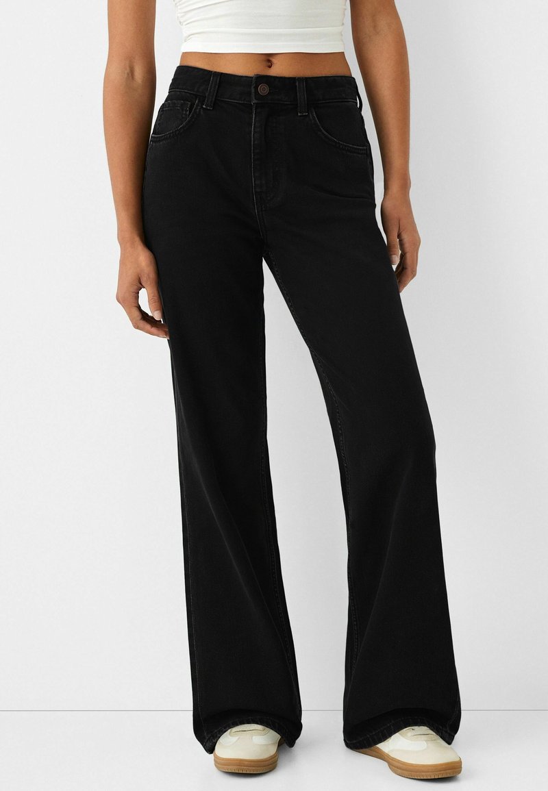 Personne portant un jean noir taille haute à jambes larges, associé à des baskets blanches et un haut court blanc sur un fond uni.