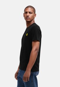 Schwarzes Baumwoll-T-Shirt mit Rundhalsausschnitt, kurzen Ärmeln und einem kleinen grünen Grafikdesign auf der linken Brust. Kombiniert mit hellblauen Jeans.