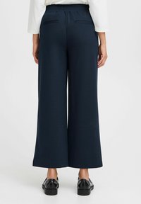 Pantaloni a gamba larga blu navy con cintura elastica, due tasche posteriori e una texture liscia, abbinati a scarpe nere.