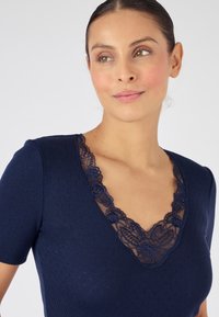 Top blu navy a maniche corte con scollatura a V, caratterizzato da dettagli di pizzo intricati attorno al collo. Tessuto testurizzato con motivi sottili.