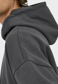Primer plano de una persona que lleva una sudadera con capucha gris oscuro, mostrando la capucha, la costura del hombro y la textura de la tela de las mangas.
