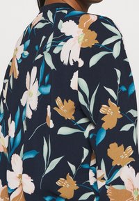 Person klädd i en marinblå klädnad med ett blommönster som har senapsgula, vita blommor och gröna blad, sedd bakifrån.