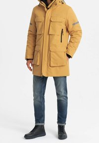 Parka amarilla acolchada con capucha, múltiples bolsillos frontales, acentos grises en las mangas y cremallera. Combinada con jeans azules y botas negras.