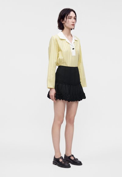 Femme debout portant un chemisier jaune à manches longues avec col blanc, mini-jupe noire à volants, et chaussures noires fermées sur fond blanc.
