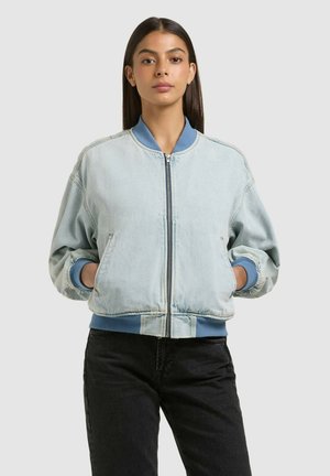 Junge Frau mit langen dunklen Haaren trägt eine hellblaue Bomberjacke mit blauen Bündchen und schwarze Hose, steht mit den Händen in den Taschen.