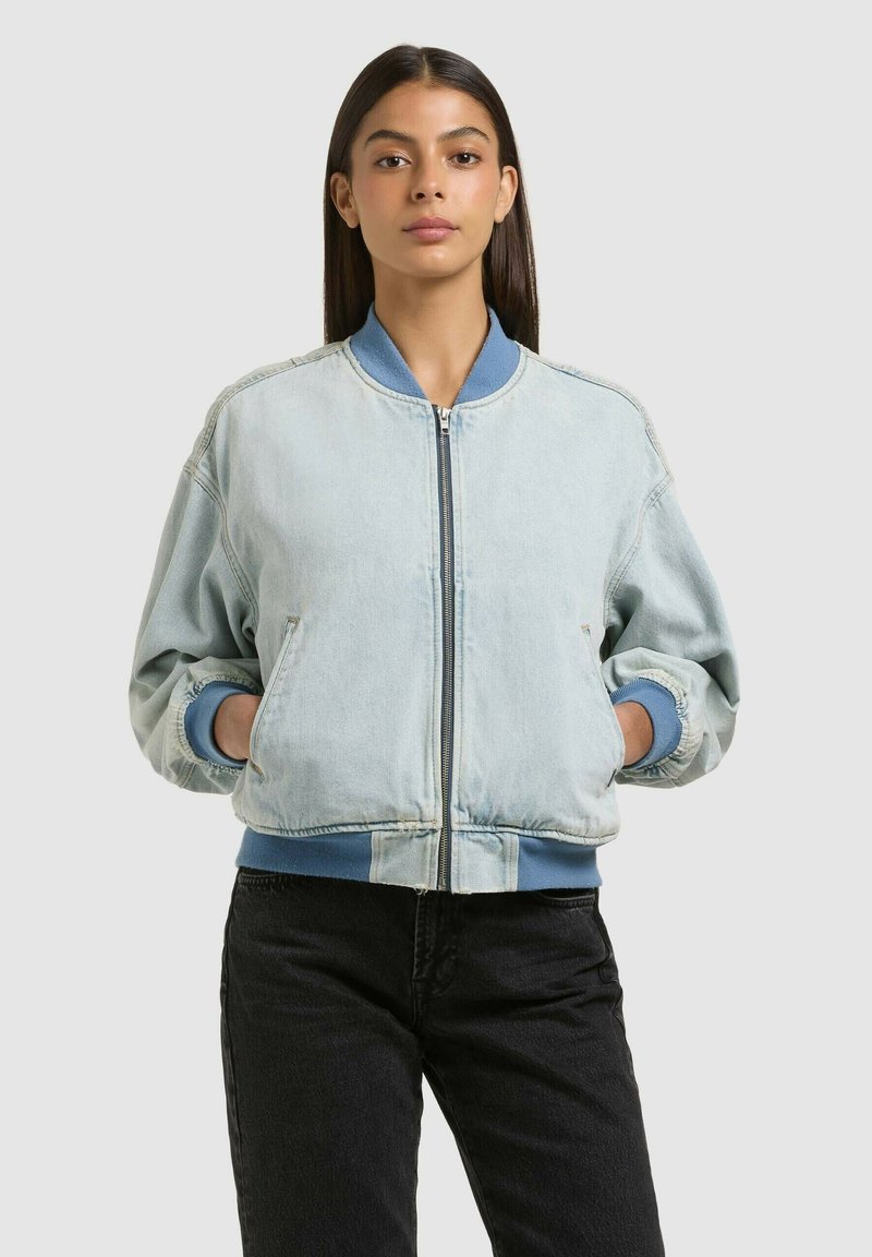 Jeune femme aux cheveux longs et foncés portant une veste légère de type bomber bleue claire avec des poignets bleus et un pantalon noir, debout les mains dans les poches.