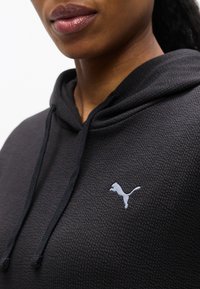 Γυναίκα που φοράει σκούρο φούτερ με υφή και μικρό λευκό λογότυπο Puma στο στήθος, εμφανιζόμενη από τους ώμους μέχρι το στόμα.