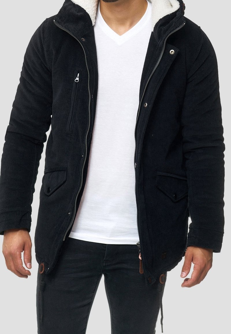Mann trägt eine schwarze Cordjacke mit Fleece-Futter über einem weißen V-Ausschnitt-T-Shirt und schwarzen Hosen vor einem schlichten hellen Hintergrund.
