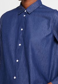 Mörkblå denimskjorta med knapp-ned krage, med vita knappar och en subtil textur. Vänster sida visar detalj av axelsöm.