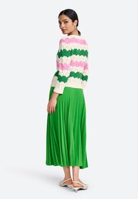 Pull en tricot avec des motifs en vagues verts, roses et crème ; associé à une jupe plissée verte fluide et des chaussures de couleur claire.