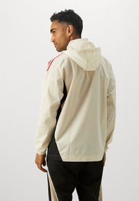 Letvægts beige jakke med hætte, sorte sidepaneler og røde detaljer på skuldrene. Har elastiske manchetter og en afslappet pasform.