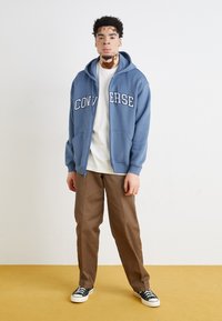 Felpa di cotone blu con il testo "CONVERSE", t-shirt bianca sotto, pantaloni marroni e sneakers black and white. Il modello è in piedi contro uno sfondo neutro.