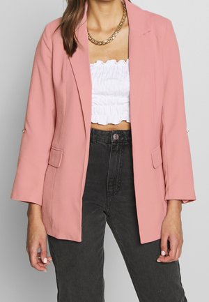 Kvinde iført en pink blazer over en hvid rynket crop top, sorte højtaljede jeans og en chunky guldkæde.