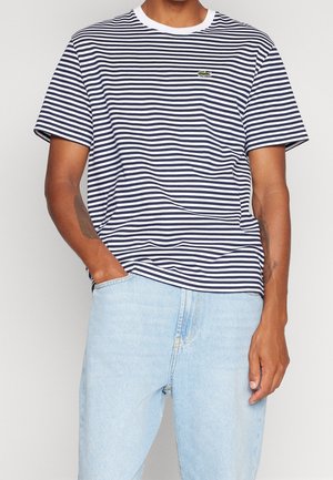 Man draagt een navyblauw-wit gestreept T-shirt met korte mouwen en een klein groen krokodillenlogo, en lichtblauwe jeans, staande met één hand in de zak.