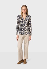 T-shirt à manches longues avec un motif abstrait bleu et beige, doté d'un décolleté en V. Accompagné de pantalons beiges et de baskets brunes.