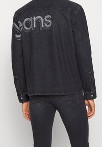 Chaqueta de denim negra con acabado mate, que presenta un gran logo gris de "jeans" en la parte posterior, botones a presión y un diseño de corte relajado.