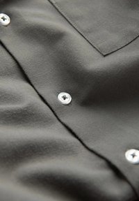 Un tissu gris foncé avec une texture lisse et un subtil éclat présente des boutons pression en métal et une poche sur la poitrine, mettant en avant son design simple.