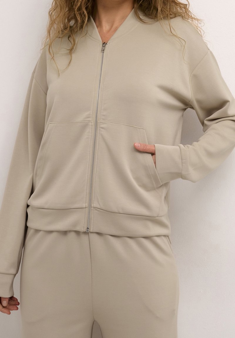 Femme portant une veste beige zippée, la main droite dans la poche avant, associée à un pantalon beige assorti.