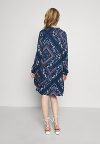 Långärmad klänning i ett mörkblått tyg med ett geometriskt mönster och accenter i teal, vinröd och vit; avslappnad passform, rynkade detaljer.