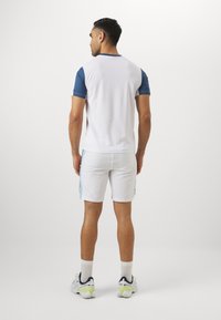 Heren sportoutfit met een witte korte mouwen top met blauwe accenten en bijpassende witte short. De stof lijkt glad en lichtgewicht.