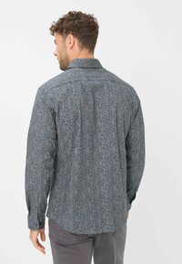 Mann, der von hinten zu sehen ist, trägt ein langärmliges, durchgeknöpftes Hemd mit Blumenmuster und graue Hose vor einem schlichten weißen Hintergrund.