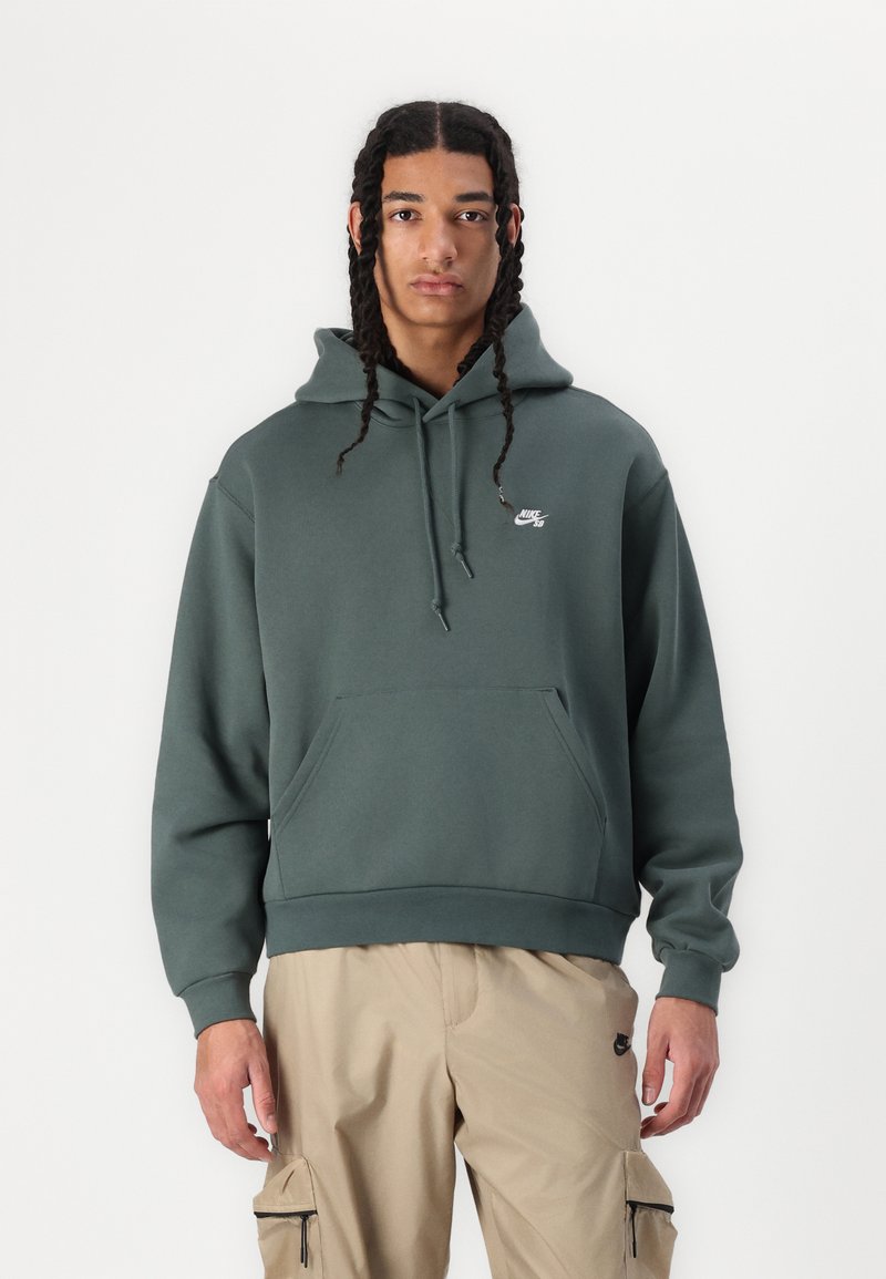 Temno zelena bombažna kapuca s sprednjo žepom, nastavljivo zategljivo kapuco in majhnim belim logotipom na prsih. Ohlapen kroj.