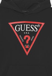 Hoodie noir avec un grand logo triangulaire rouge comportant un texte blanc, y compris "GUESS", "U.S.A." et des chiffres, ainsi qu'une poche frontale.