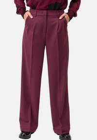 Pantaloni larghi color bordeaux con pieghe frontali, una vita e tasche laterali, realizzati in materiale liscio. Abbinati a scarpe nere per ricreare un contrasto.