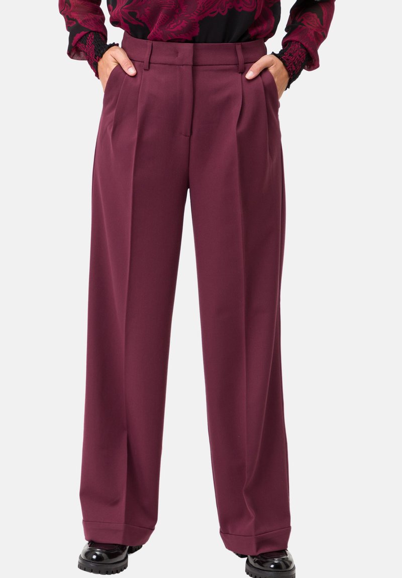 Pantaloni larghi color bordeaux con pieghe frontali, una vita e tasche laterali, realizzati in materiale liscio. Abbinati a scarpe nere per ricreare un contrasto.