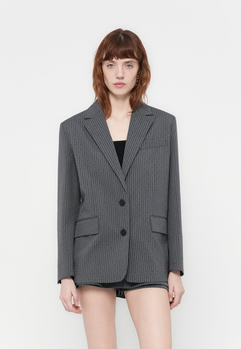Eine Frau mit roten Haaren trägt ein graues Oversized-Blazer mit Nadelstreifen und passende Shorts und steht vor einem schlichten weißen Hintergrund.