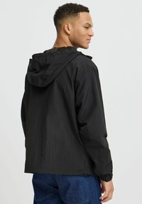 Veste à capuche noire en tissu léger avec une texture lisse, des poignets élastiques et une coupe décontractée, associée à un pantalon en denim bleu.