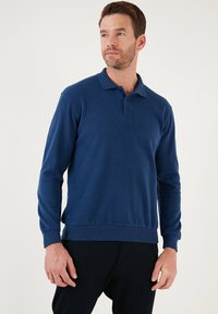Tengerészkék, hosszú ujjas pólóing, bordás anyagból, hajtott gallérral, három gombos záródással és formázott aljjal. A modellen sötét nadrág van.