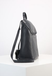 Sac à dos en cuir noir au design épuré, avec un dessus zippé, une bandoulière réglable et une texture lisse. Accents minimalistes présents.