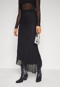 Falda midi de encaje negro con dobladillo rizado y patrón texturizado, combinada con botas de cowboy negras con acentos plateados y un pequeño bolso plateado.