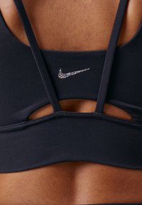 Crna atletska grudnjak s tankim trakama i izrezima. Ima mali, sjajni Nike logo na stražnjoj strani, izrađen od glatkog materijala.
