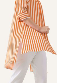 Tunic a righe arancione e bianca con orlo asimmetrico e spacchi laterali. Tessuto morbido che mostra righe verticali, con maniche arrotolate e vestibilità comoda.