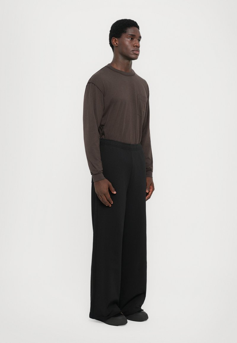 Een bruine lange mouwen shirt met een zak, gecombineerd met losse zwarte broek. De stof lijkt glad, met een relaxte pasvorm en een uniforme kleur.