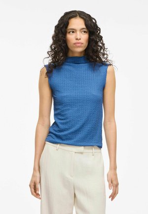 Top senza maniche in un tessuto blu testurizzato con un collo alto, abbinato a pantaloni leggeri e larghi.