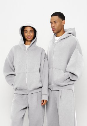 Dos personas que llevan sudaderas con capucha oversize de color gris claro con cremallera y pantalones a juego están de pie sobre un fondo claro y liso.
