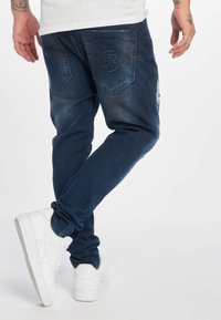 Sötétkék farmernadrág texturált felülettel, szűk�ülő lábszárakkal és kopott foltokkal a combon, hímzett hátsó zsebekkel.