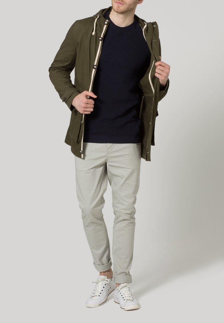Veste vert olive avec capuche, cordons de serrage crème, pull bleu marine, pantalon gris clair et baskets blanches. Tenue décontractée et superposée.