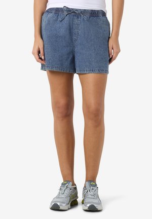 Kvinne iført blå denimshorts med elastisk midje og knyting foran, kombinert med grå og gule løpesko, stående foran en hvit bakgrunn.
