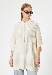 Poncho de punto color crema con capucha, que presenta una textura acanalada y mangas anchas. Combinado con unos jeans de mezclilla azul claro.
