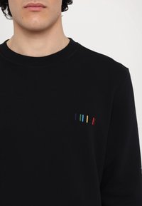 Gros plan d'une personne portant un sweat-shirt noir avec de petites rayures verticales brodées en bleu, sarcelle, jaune et rouge sur la poitrine gauche.