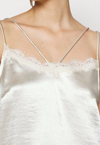 Silky silver camisole featuring delicate lace trim along the neckline, spaghetti straps, and a smooth texture. Close-up view.