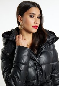 Sort puffersjakke med en blank finish, udstyret med en pelsforet hætte, store knapper og en beltestrop om taljen. Modellen har tilføjet guldøreringe som accessory.